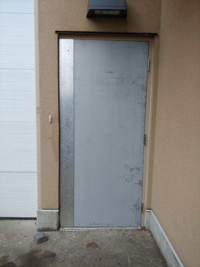 A flat gray door with a metallic frame, set against a textured tan wall.