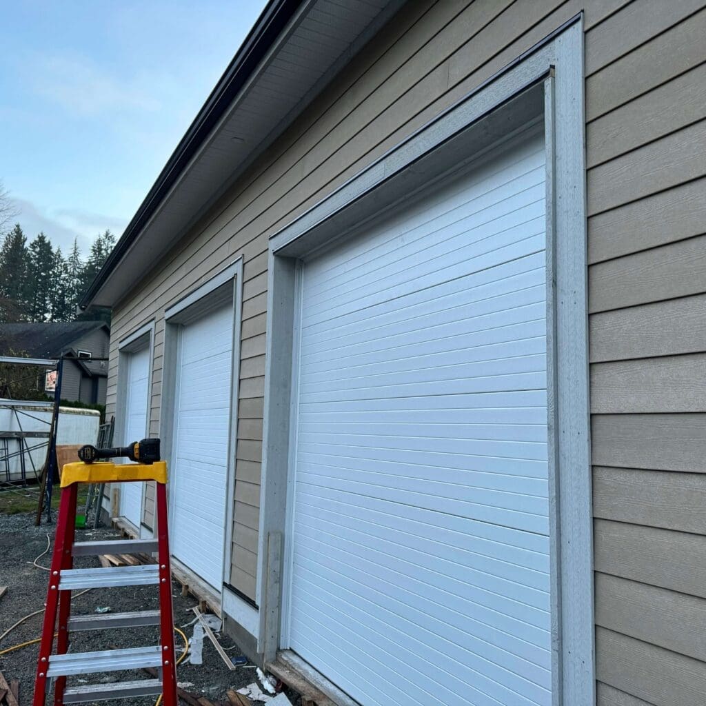 A partially constructed building exterior featuring two white garage doors and a ladder in front.