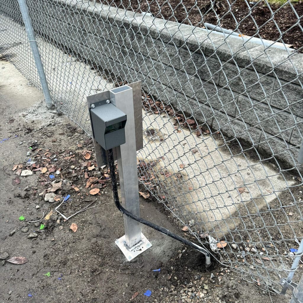 A metal pole with a small green utility box attached to it, standing near a chain-link fence.