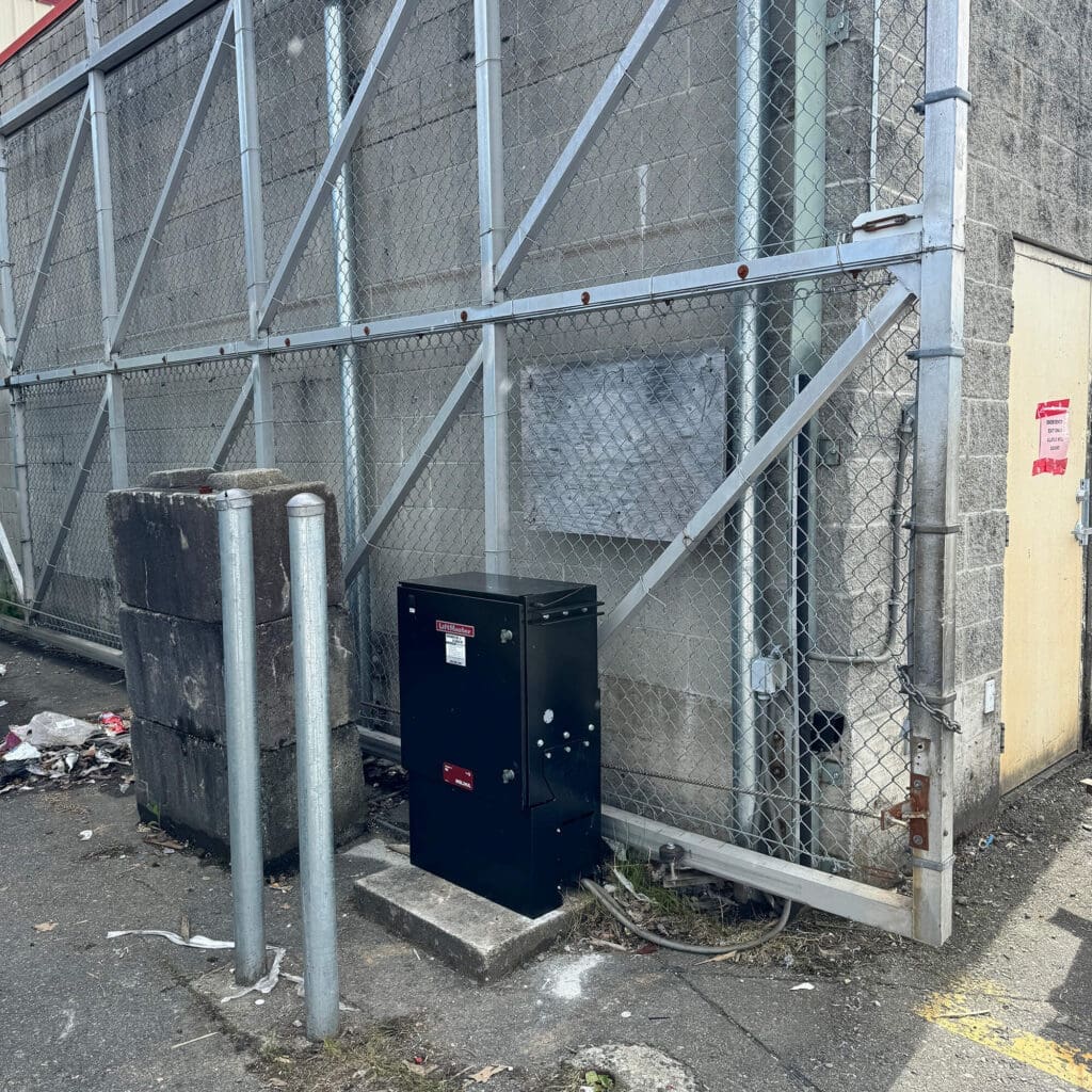 Black electrical box against a gray wall, surrounded by a chain-link fence.