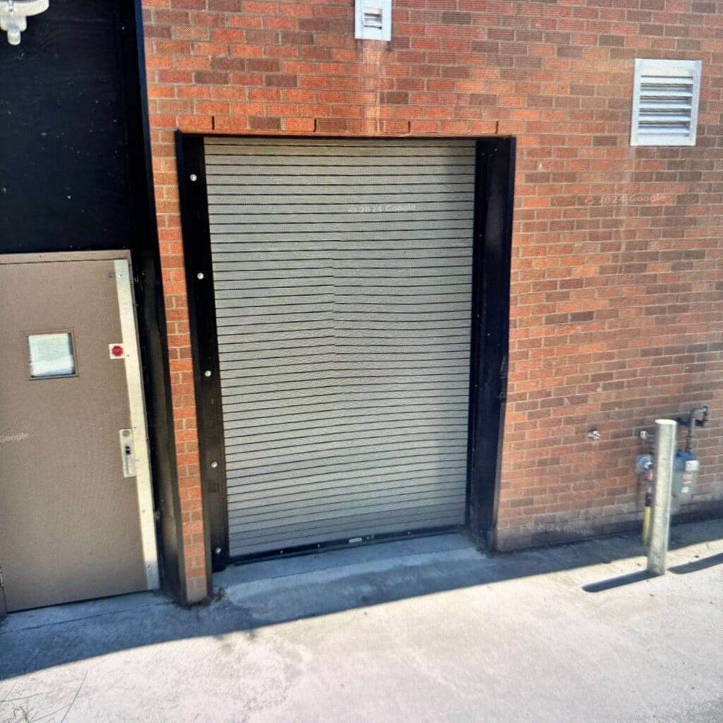 A closed metal roll-up door on a brick wall, with a small door beside it and utility pipes visible on the right side