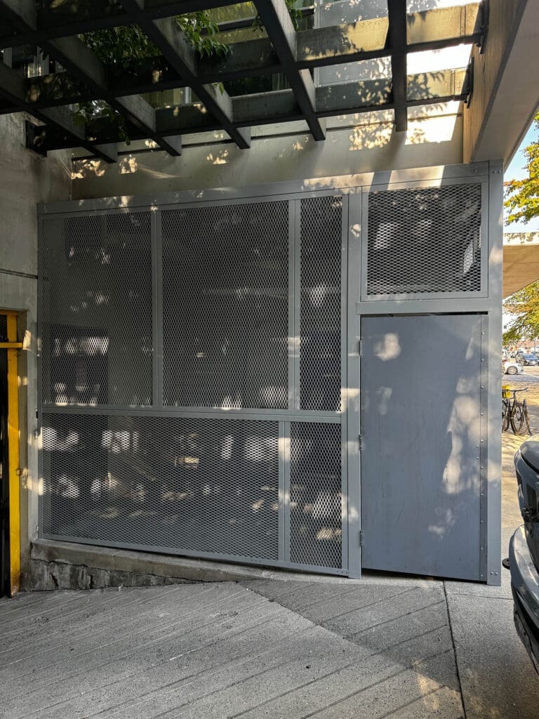 A gray metal gate with a mesh pattern is mounted on a wall, partially obscured by shadows from overhead foliage.