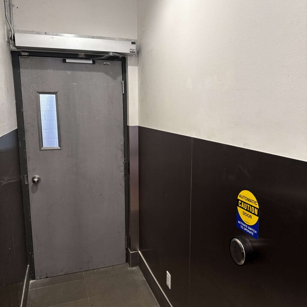 A door with a small window is seen at the end of a corridor that features dark walls.