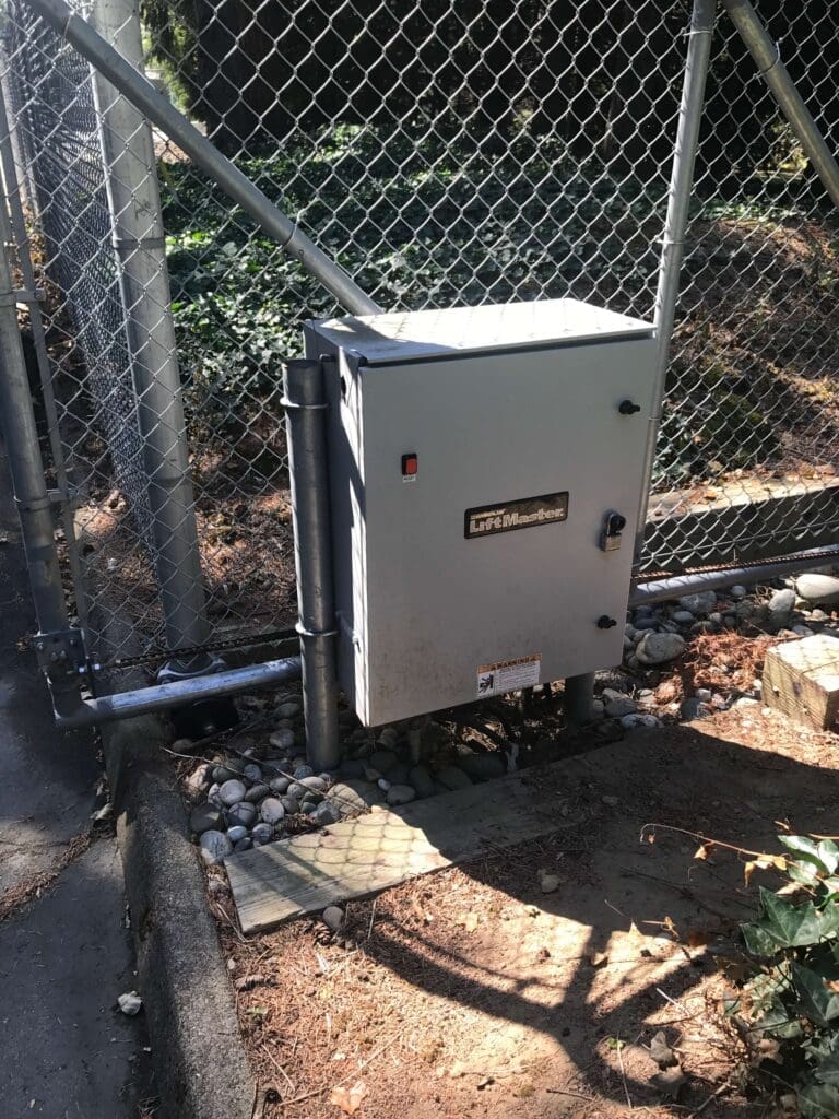 A gray electrical control box mounted on a fence, surrounded by rocks and greenery.