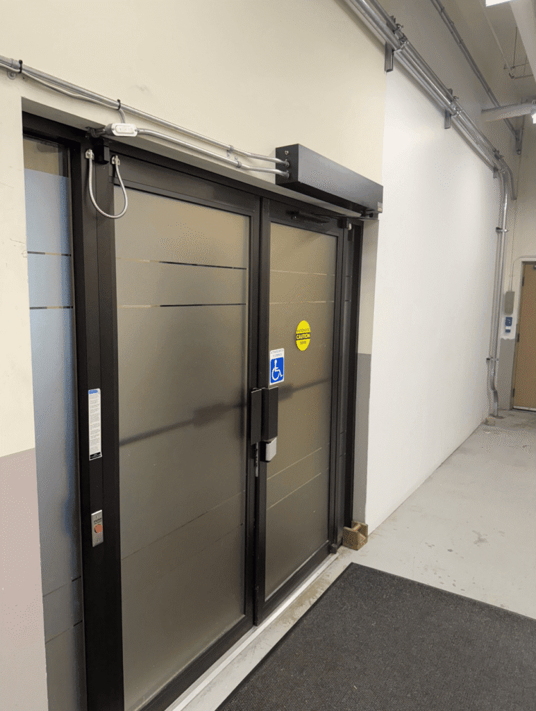 Image of a glass double door with a push-button automatic opener, located in a corridor.