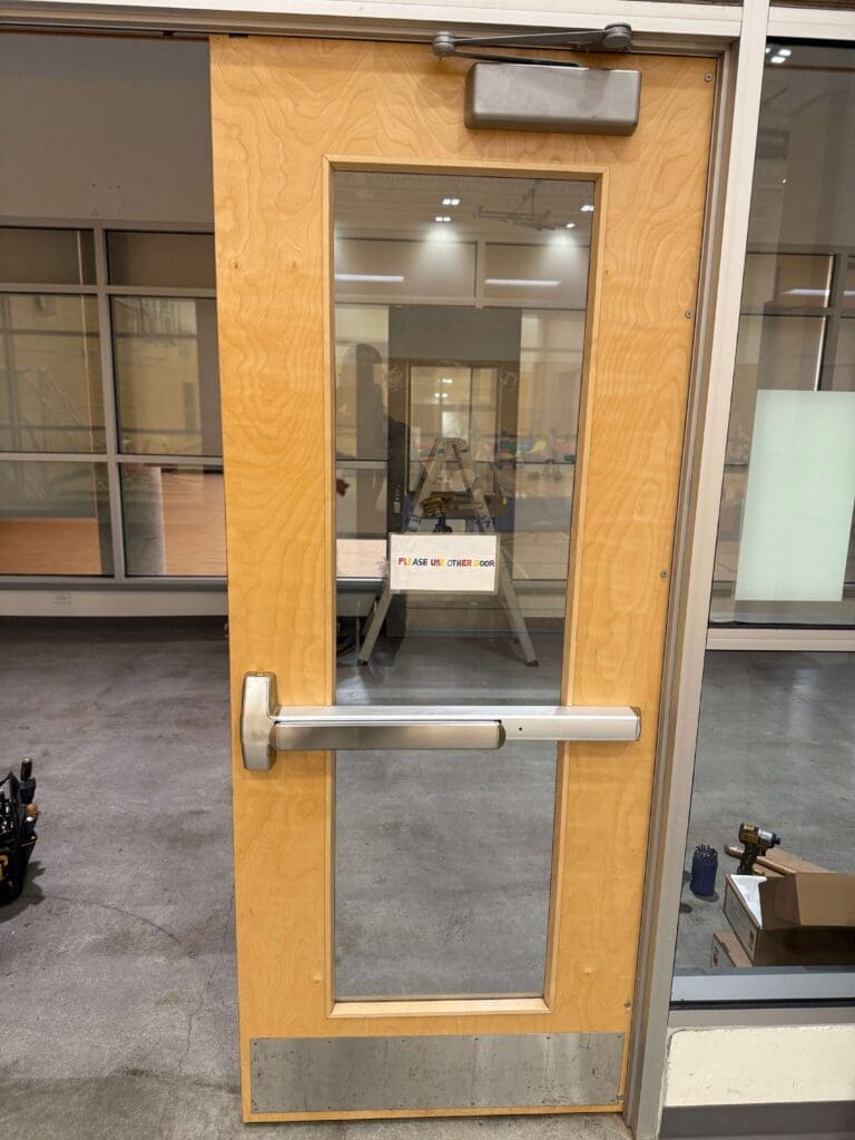 A wooden door with a large rectangular glass window, featuring a metal push bar handle, set in a modern indoor environment with a gray floor.