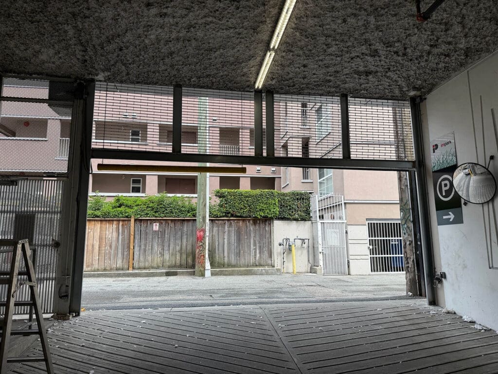 View of an open garage with a concrete floor, showing a street outside and a wooden fence in the background.