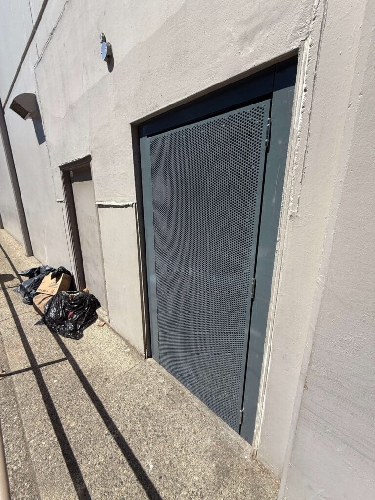 A gray door with a wire mesh pattern, set in a plain wall, next to a pile of black garbage bags on a concrete surface.