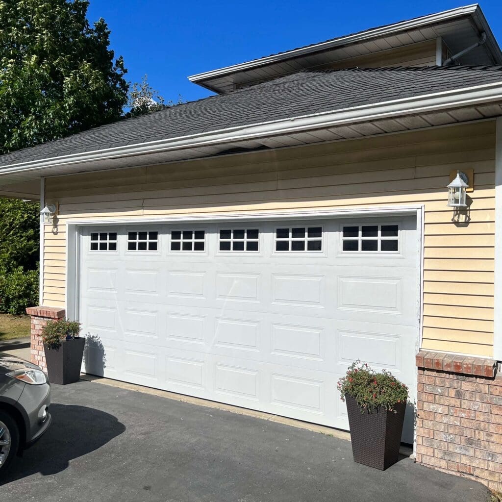 A residential garage with a white door featuring small windows at the top, surrounded by a landscaped area and a parked car.