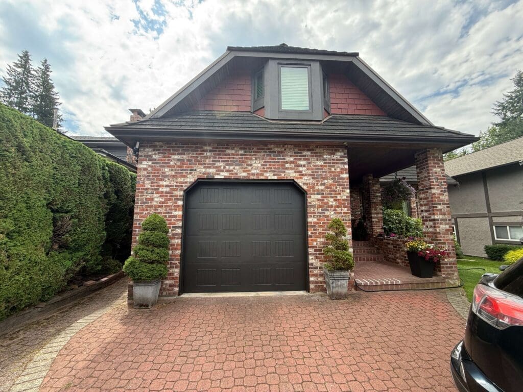 A brick house with a gabled roof and a black garage door, surrounded by neatly trimmed hedges and potted plants on a brick driveway