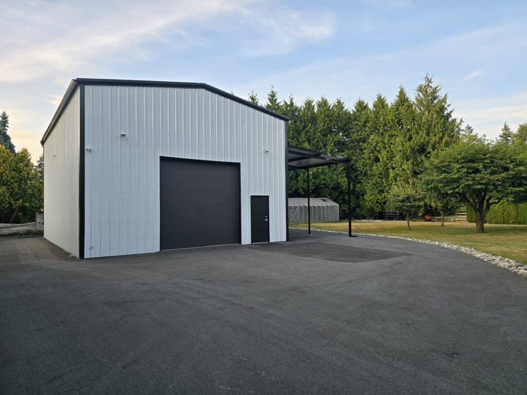 A large, modern white metal building with a black garage door, situated on a paved area surrounded by green trees and shrubs.