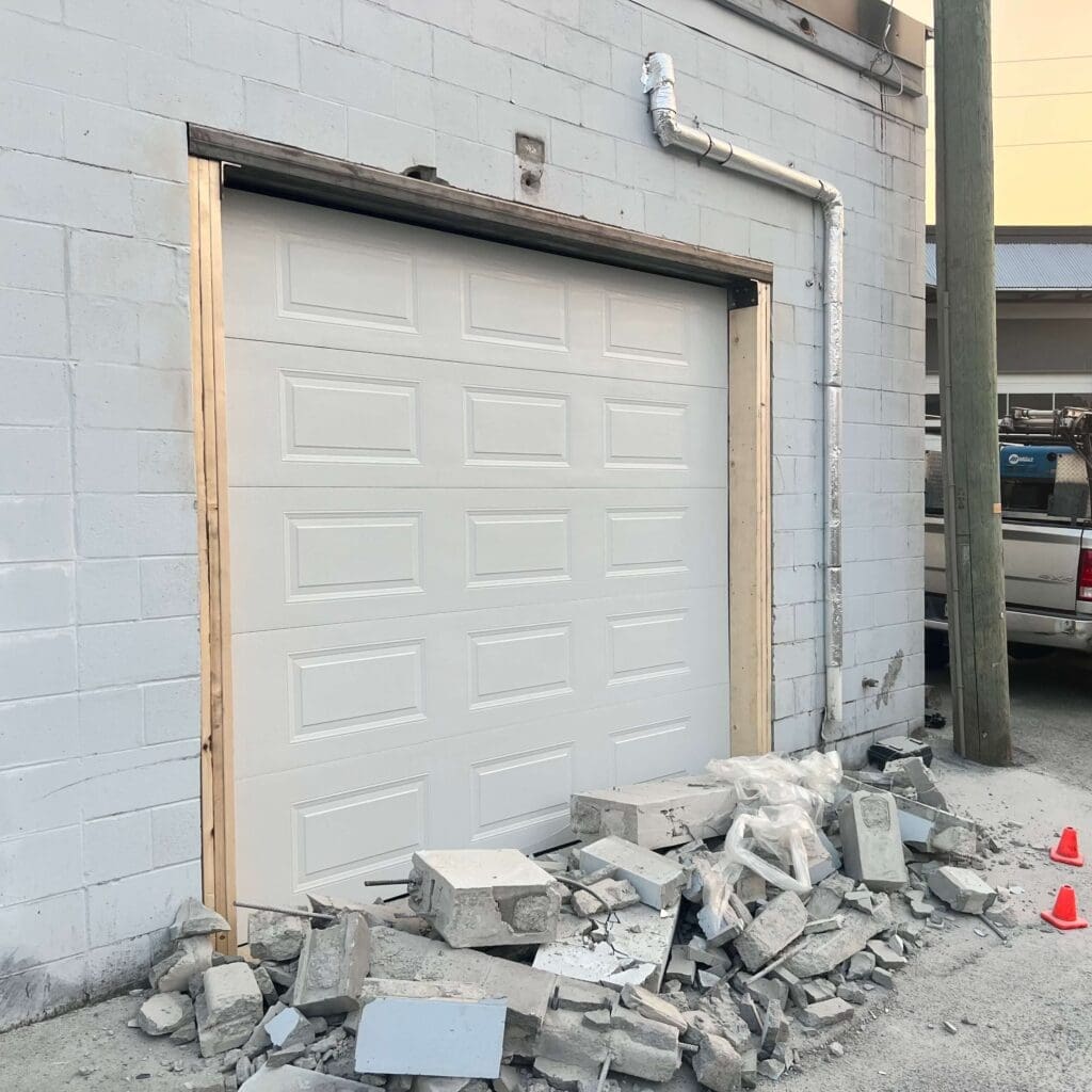A partially constructed garage door frame with a white door installed, surrounded by debris from broken concrete blocks on the ground