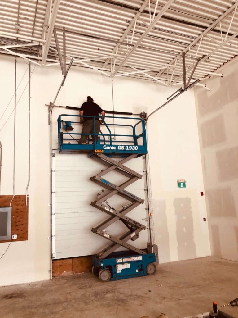 A person working on the garage door of a warehouse or industrial space using a blue scissor lift.