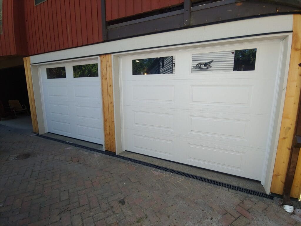 Two white garage doors with windows, set against a wooden structure and a paved driveway.